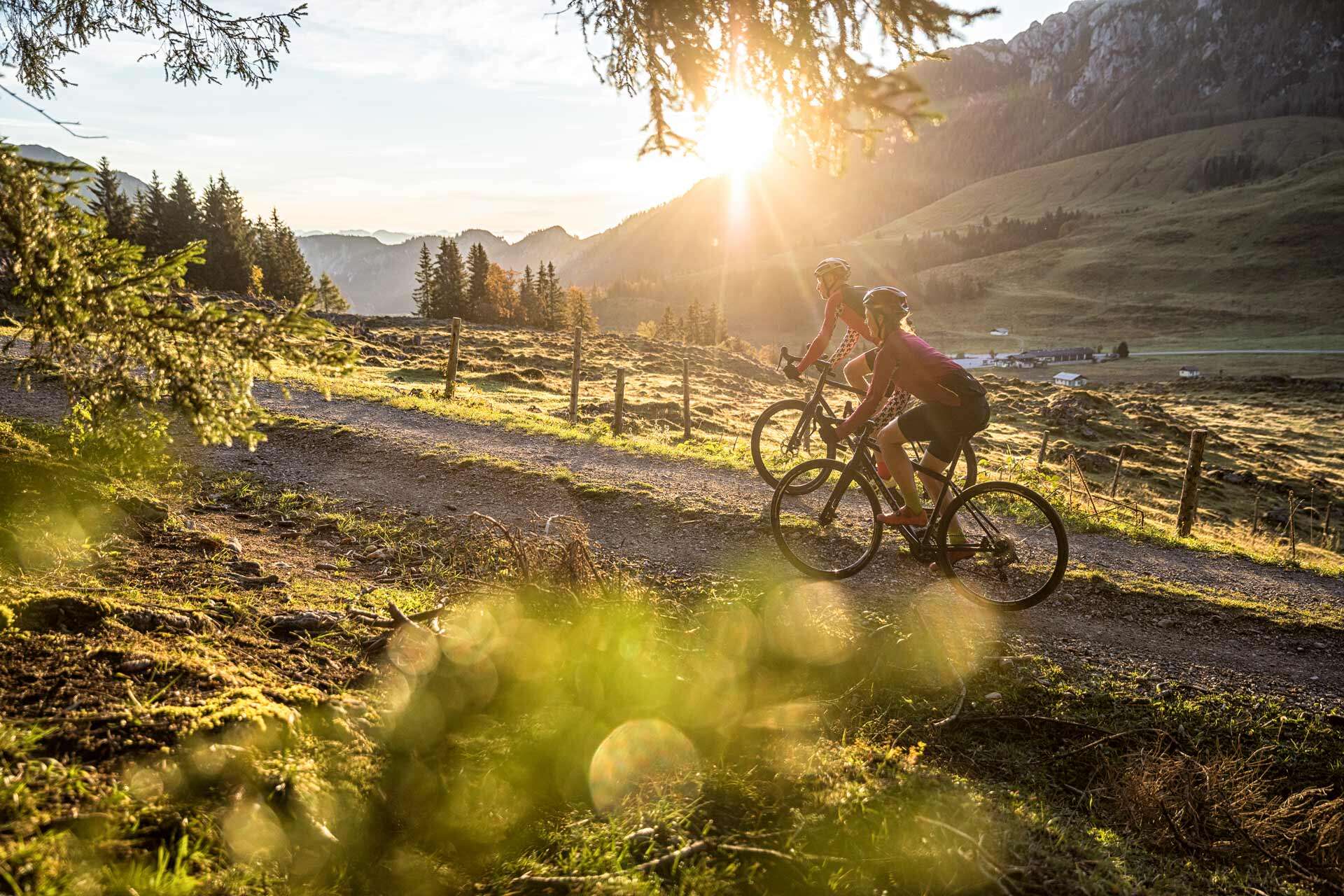 Zwei Radfahrerinnen auf einem Schotterweg durch die Almlandschaft bei Gegenlicht